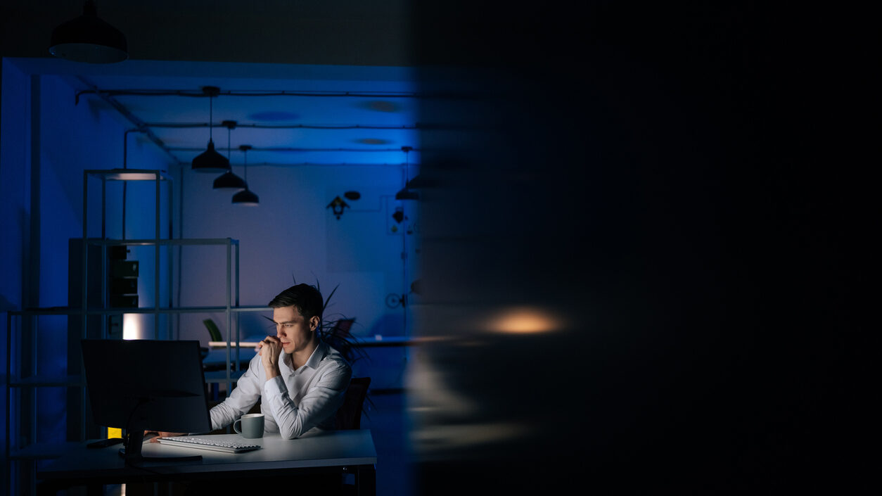 Remote view of serious young business man remote working overtime, learning online late at night in in dark room with neon light using desktop computer at workplace.