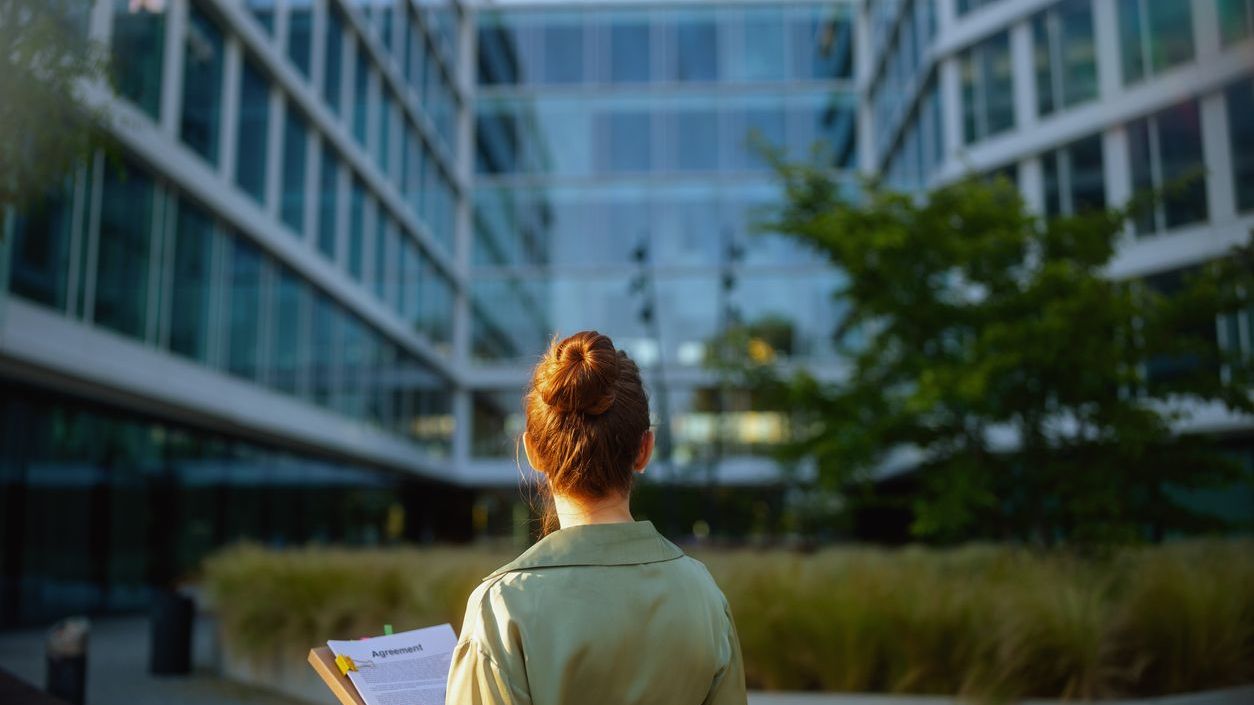 Seen from behind business woman near office building in green blouse with documents.