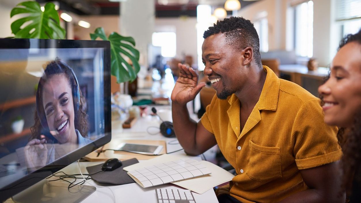 A Black man waves to his colleague on a video call from his office